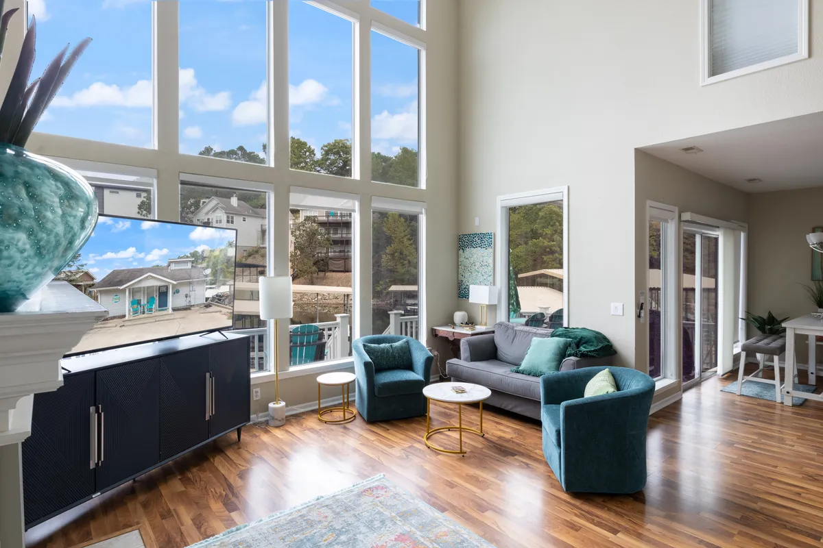 The image presents a bright, inviting living space with large windows letting in abundant natural light, comfortable seating arranged for conversation or relaxation, and a television. A decorative vase adds a colorful accent, and the overall mood is cozy and welcoming.