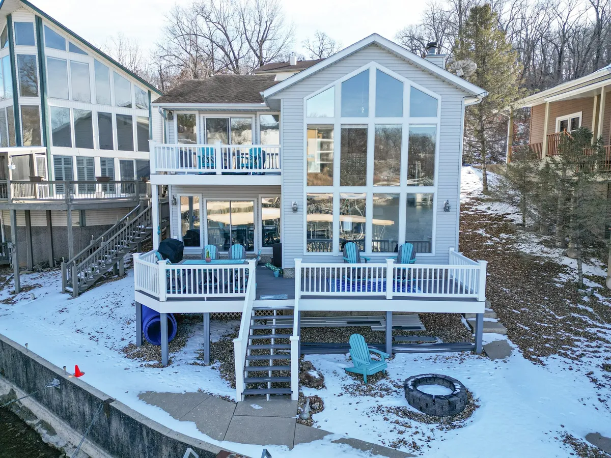 A spacious lakeside deck featuring large glass windows, multiple chairs arranged around a modern fire pit, and views out to the water. The cozy setup encourages gatherings, storytelling, and stargazing under the open sky.