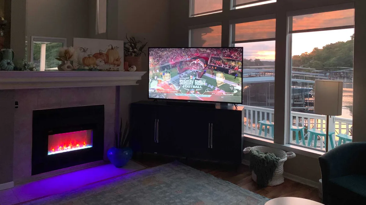 This image showcases a casual and comfortable living room featuring a television displaying a sports event, an electric fireplace with glowing lights, and large windows that offer a beautiful view of the water. Decorative elements adorn the mantle, and the sunset outside bathes the space in warm light.