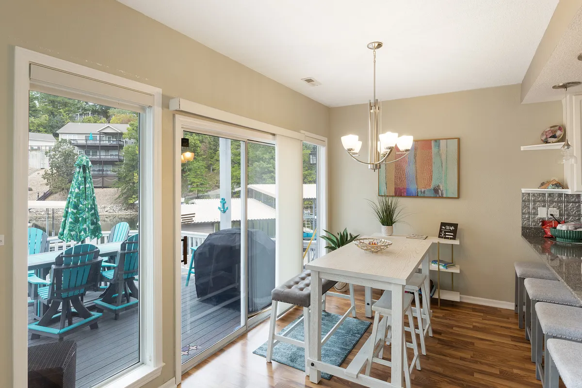 A welcoming dining area featuring a well-set dining table, bar stools, a chandelier, large sliding glass doors, and a colorful painting. The doors open directly onto the deck, blending indoor and outdoor dining possibilities.