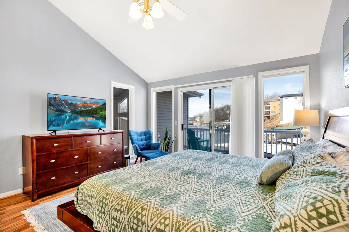 A spacious master bedroom featuring a king-sized bed with patterned bedding, large windows that fill the room with natural light, a flat-screen TV on the dresser, and a comfortable blue armchair. The windows offer glimpses of the outdoors, hinting at serene lake views.