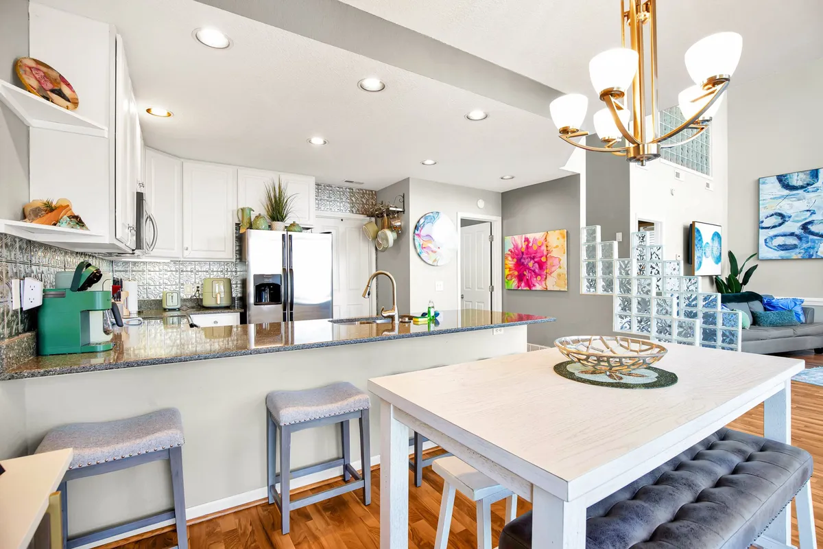 This photo captures the adjacent dining and kitchen area, featuring a stylish kitchen island, a chandelier above, a glass block wall, vibrant artwork, and a white dining table. The space is set for socializing, cooking, and entertaining, with an open-concept layout connecting it to the living area.