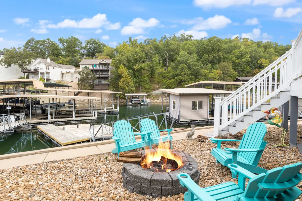 The image features the exterior of a lakefront single-family home in Osage Beach, Missouri. In the foreground, a circle of Adirondack chairs surrounds a crackling fire pit, set on a stone patio overlooking the tranquil water. The boathouse sits nearby, and a beautiful sunset glows over the lake, inviting guests to imagine evenings spent by the fire and days filled with boating and fishing adventures.