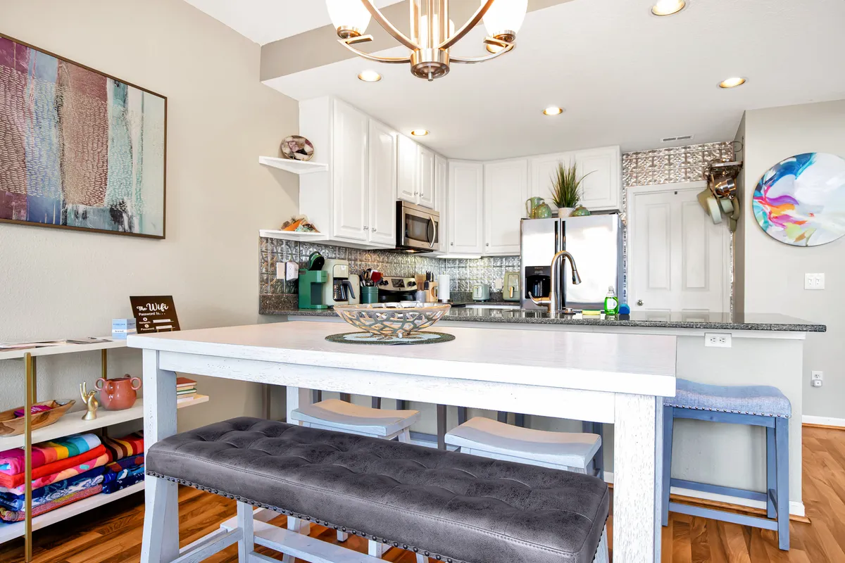A bright, modern kitchen with a central island, colorful seating cushions, modern light fixture, and a decorative art piece on the wall. The space is open, inviting, and clearly designed for both meal preparation and casual gatherings.