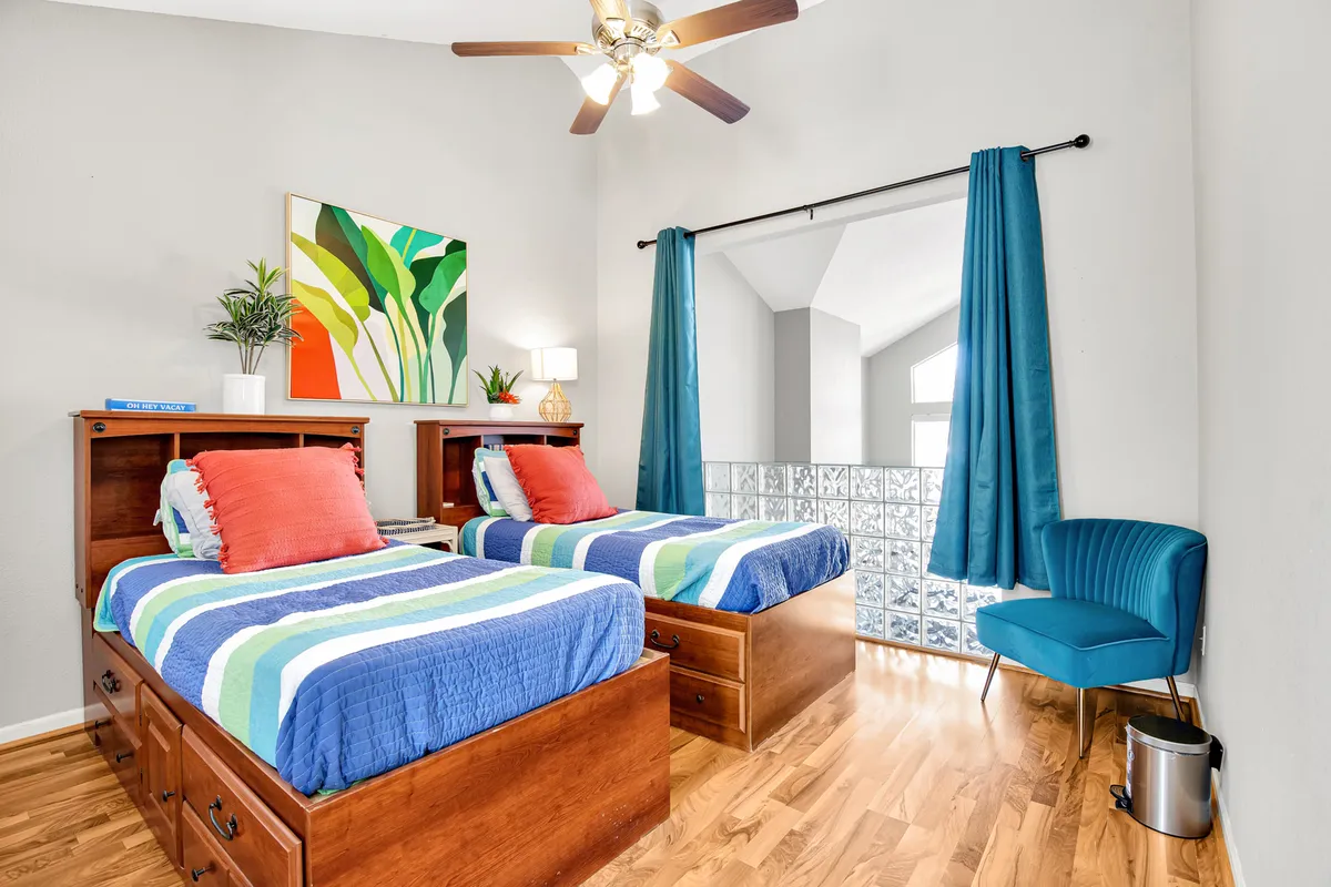 A cheerful guest bedroom with two twin beds featuring colorful bedding, decorative wall art, a cozy blue armchair, and a glass block window allowing in soft, natural light. The playful décor speaks to comfort for families or groups.