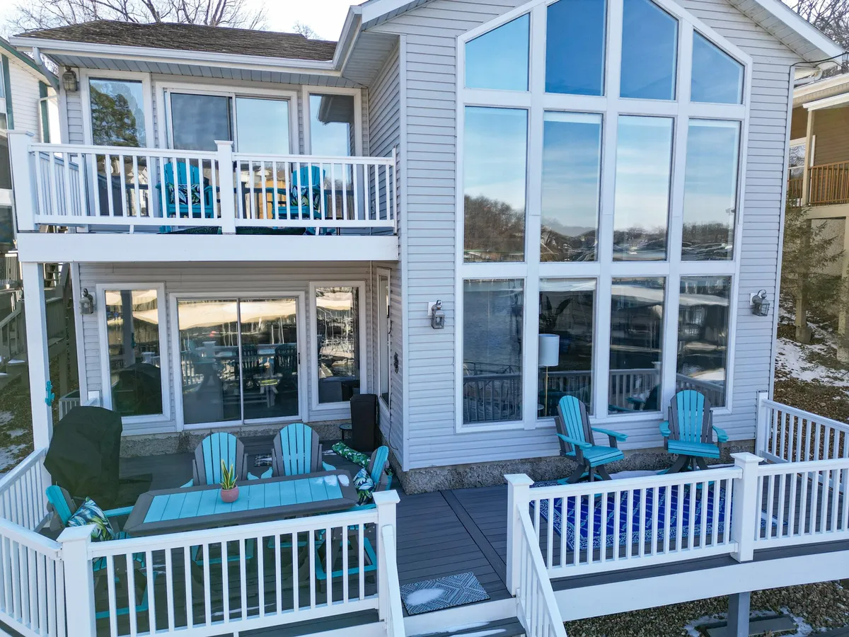 An exterior view of the house showcases large windows, colorful LuxCraft outdoor furniture, and upper-level balconies overlooking the lake. The scene is vibrant and inviting, hinting at both comfort and spectacular waterfront views from within and outside the home.