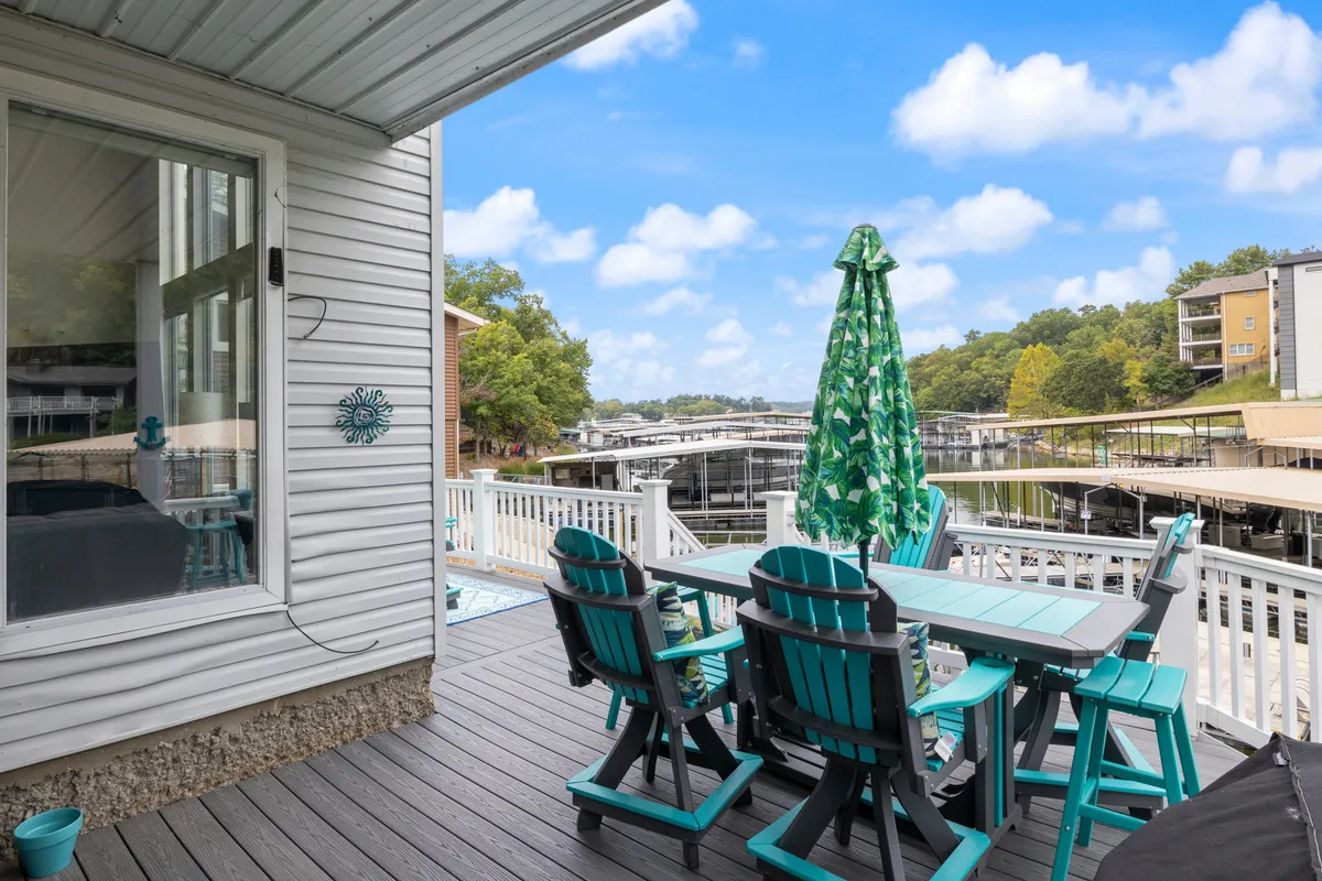An inviting outdoor dining area featuring a table with chairs beneath a green patterned umbrella, with views of the marina and the lakeside in the background. The space is set up for social gatherings and overlooks the water, suggesting both relaxation and lively group activities.